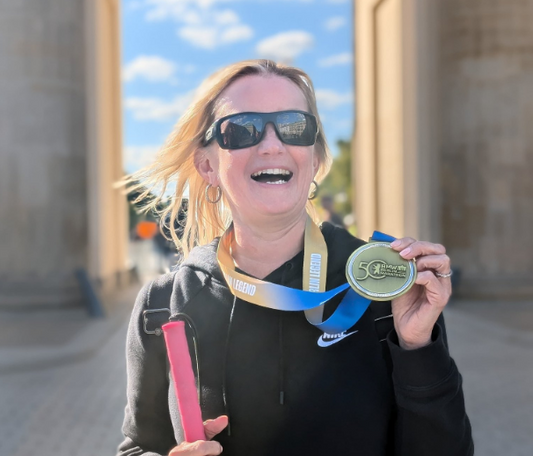 Kelly Barton, a white blonde female, wearing sunglasses, holding a pink cane in one hand and a runners medal in the other. Kelly has a big smile.