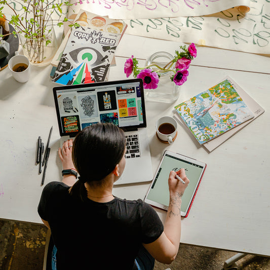 Image is looking down at the back of a woman who is working on a laptop with magazines and drinks around her