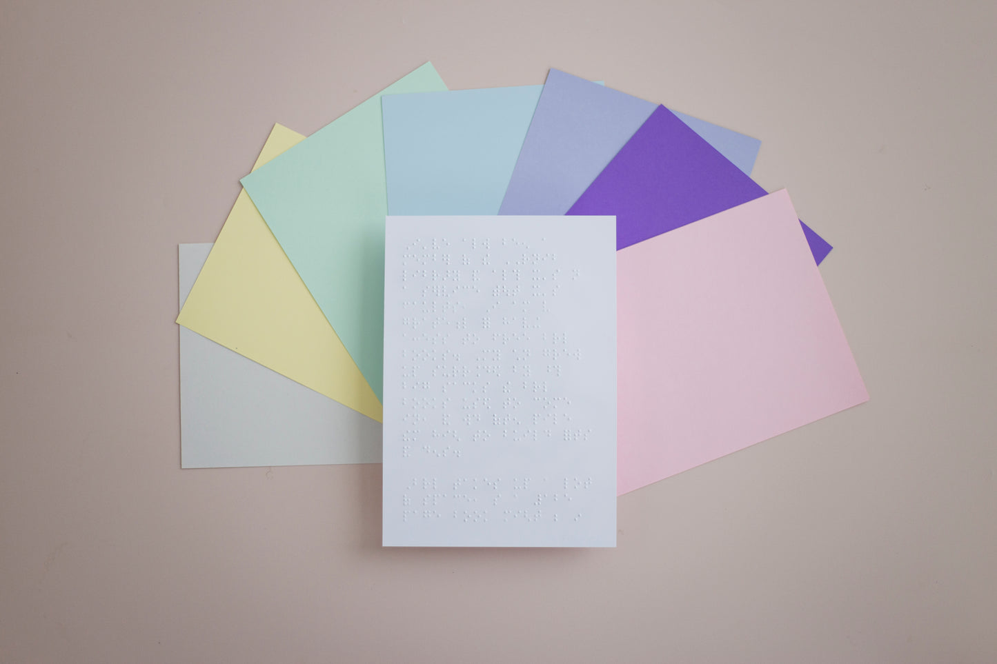 A white Braille document on top of A5 sheets in pastel green, yellow, blue, purple, pink, vibrant purple and off natural white, fanned out on a beige surface.