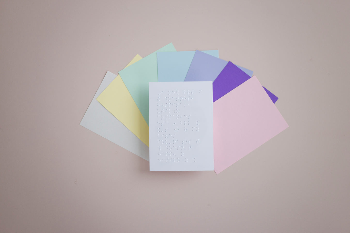 A white Braille document on top of A5 sheets in pastel green, yellow, blue, purple, pink, vibrant purple and off natural white, fanned out on a beige surface.