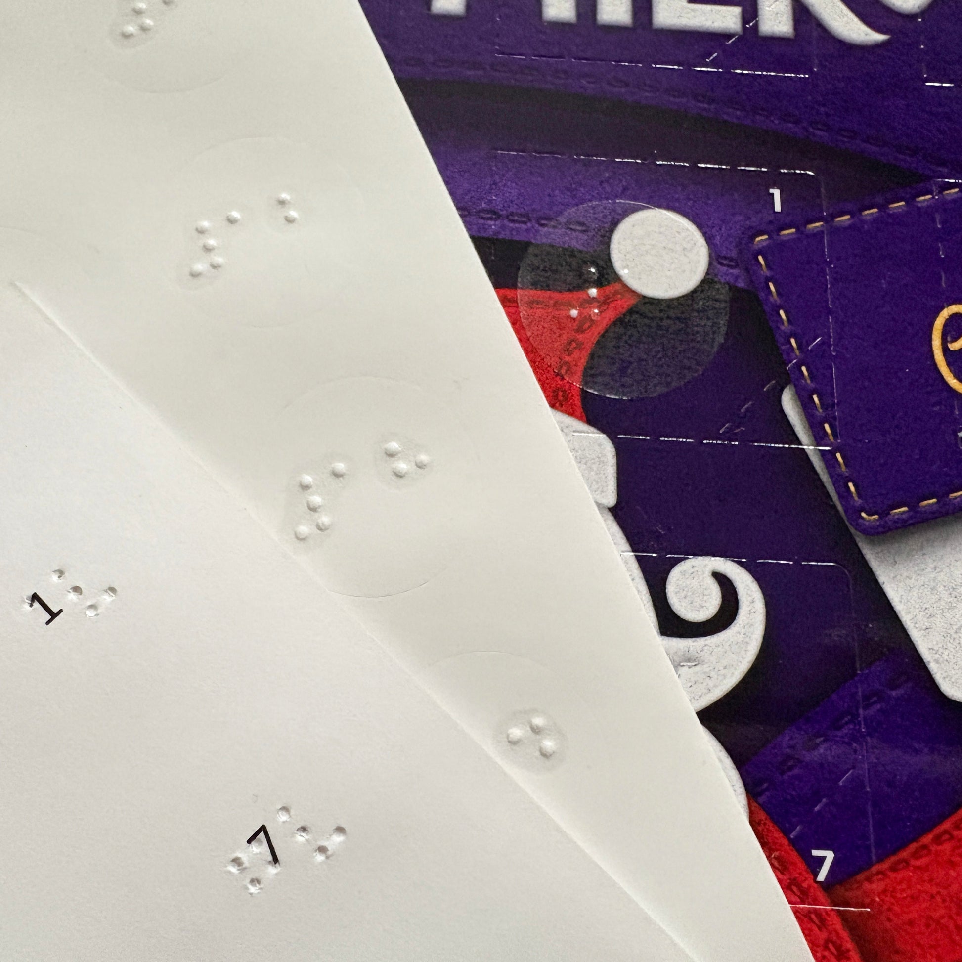 Close up of an advent calendar with the number 1 door showing a braille sticker. A sheet of braille labels is visible, along with a sheet showing the back of the numbers, which has the printed text.