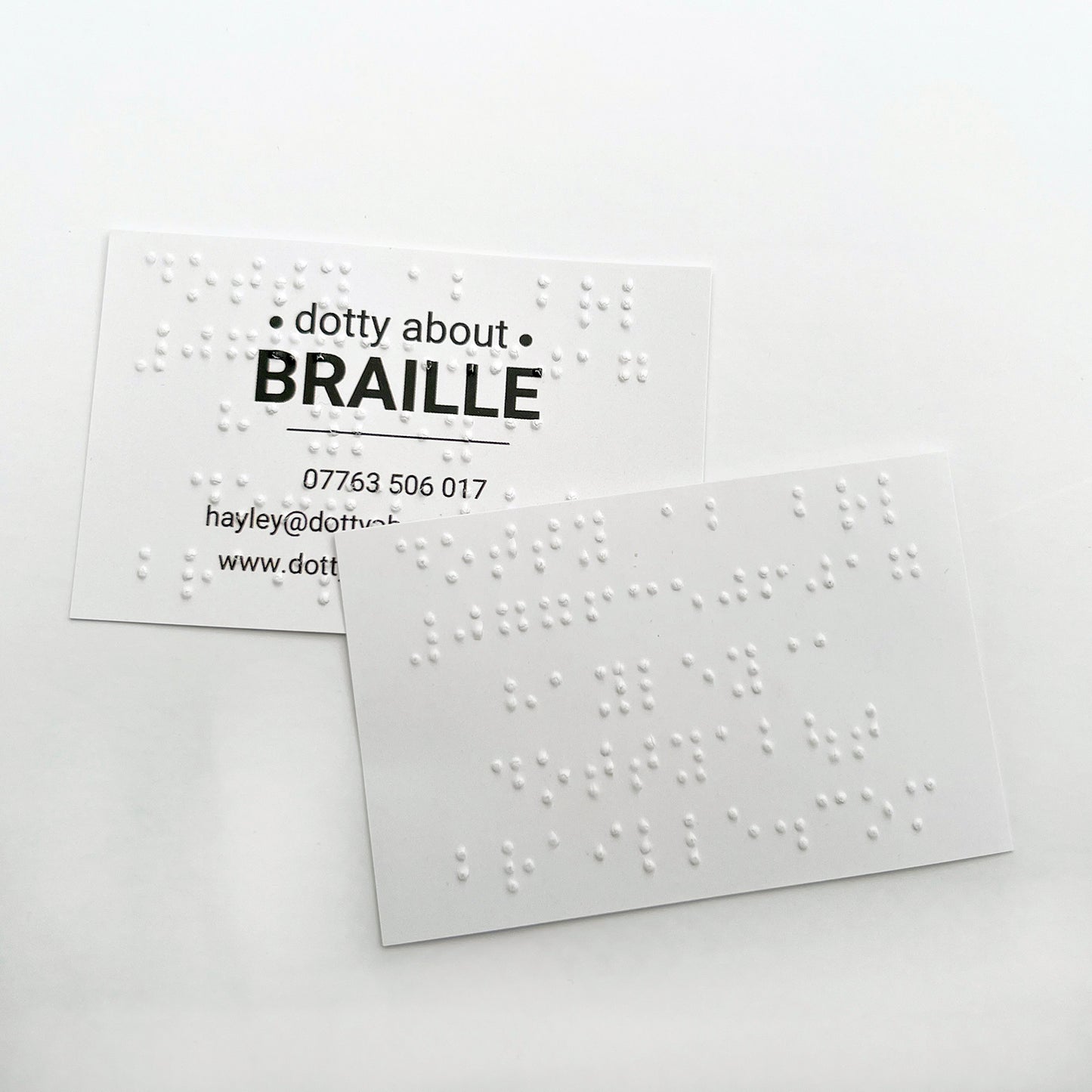 Two Dotty About Braille business cards, one has the braille on the front of the card, overlaid on the printed text, the other has the braille printed on the reverse of the business card.