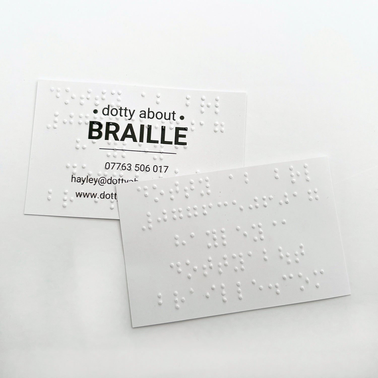 Two Dotty About Braille business cards, one has the braille on the front of the card, overlaid on the printed text, the other has the braille printed on the reverse of the business card.