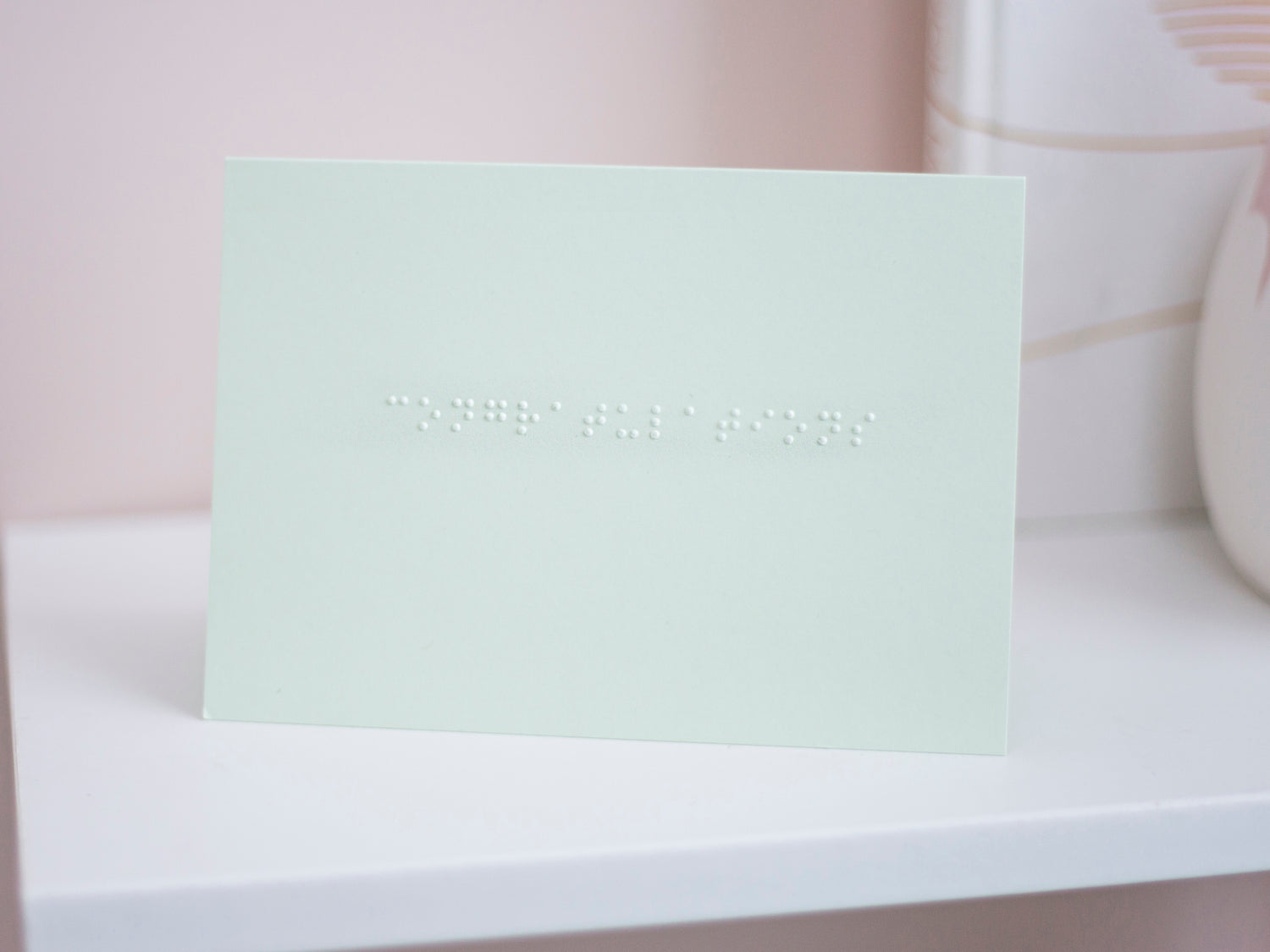 A pastel green card with congratulations written in lower case braille. The card is sat on a white shelf.