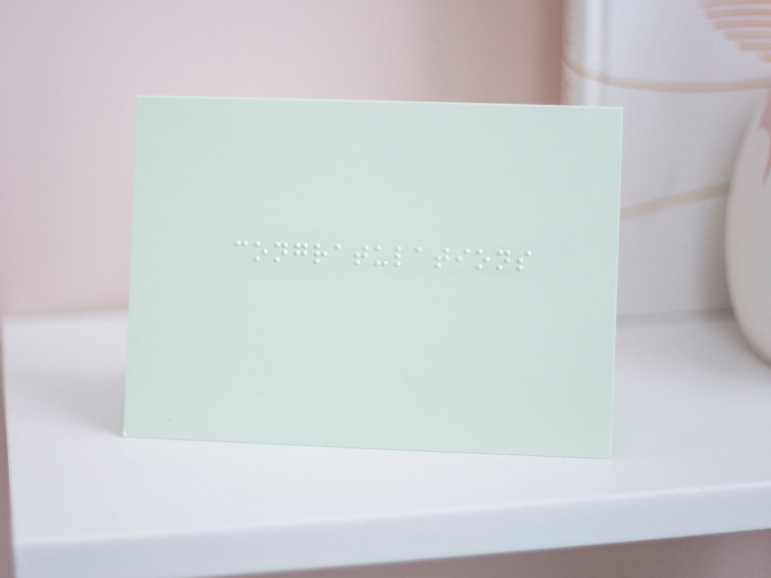 A pastel green card with congratulations written in lower case braille. The card is sat on a white shelf.