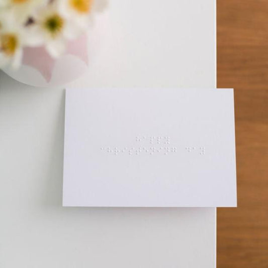 A white greetings card with happy christening day written in lower case braille. There is a blurry vase of flowers at the top of the image.