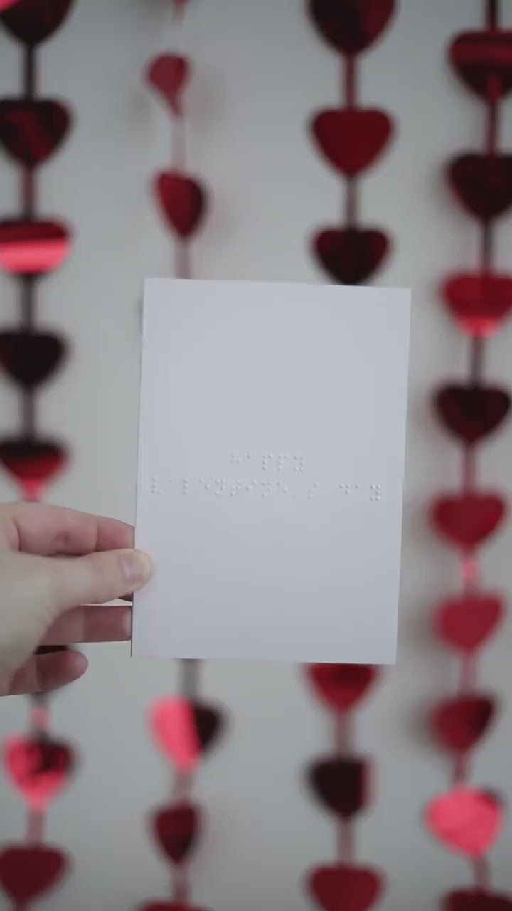 A video of a pale pink card with Happy Valentine's Day written in braille. There are hanging red shiny love hearts in the background.