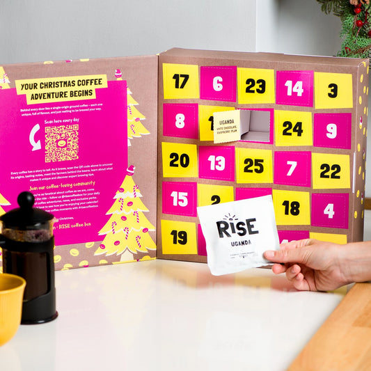 Open RISE Coffee Advent Calendar showing numbered doors and a sachet of Uganda coffee held in hand beside a mug and cafetière.