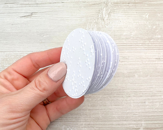 A hand wearing pink nail varnish holding small 5cm circles of white braille paper.