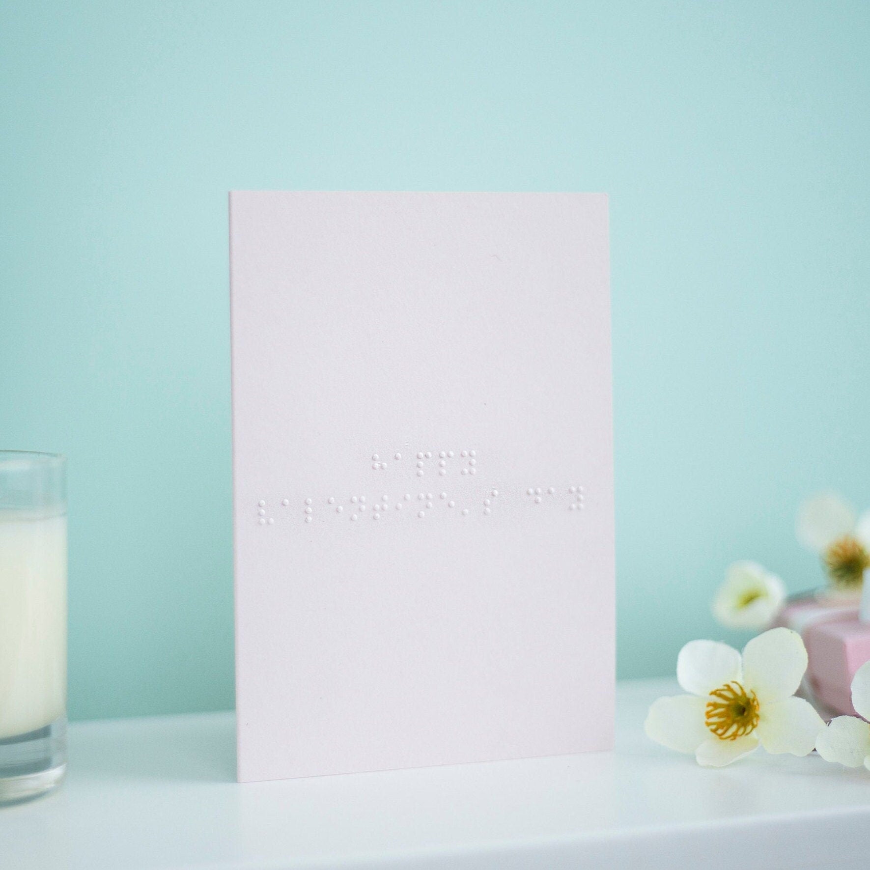 A pastel pink card with Happy Valentine's Day written in lower case braille. There is a candle to the left and a flowers to the right.