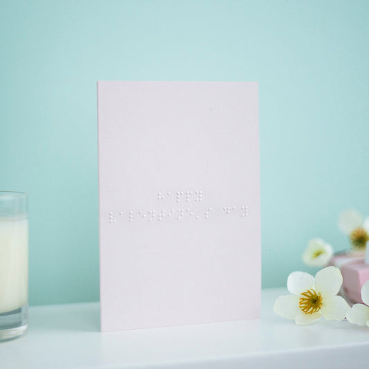 A pastel pink card with Happy Valentine's Day written in lower case braille. There is a candle to the left and a flowers to the right.