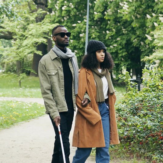 A male blind person of colour being guided by a female person of colour. The man is holding a white cane.