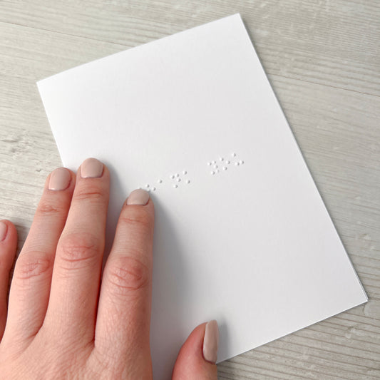 A white braille card with thank you written in lower case braille, a hand wearing pink nail varnish is on top of the card