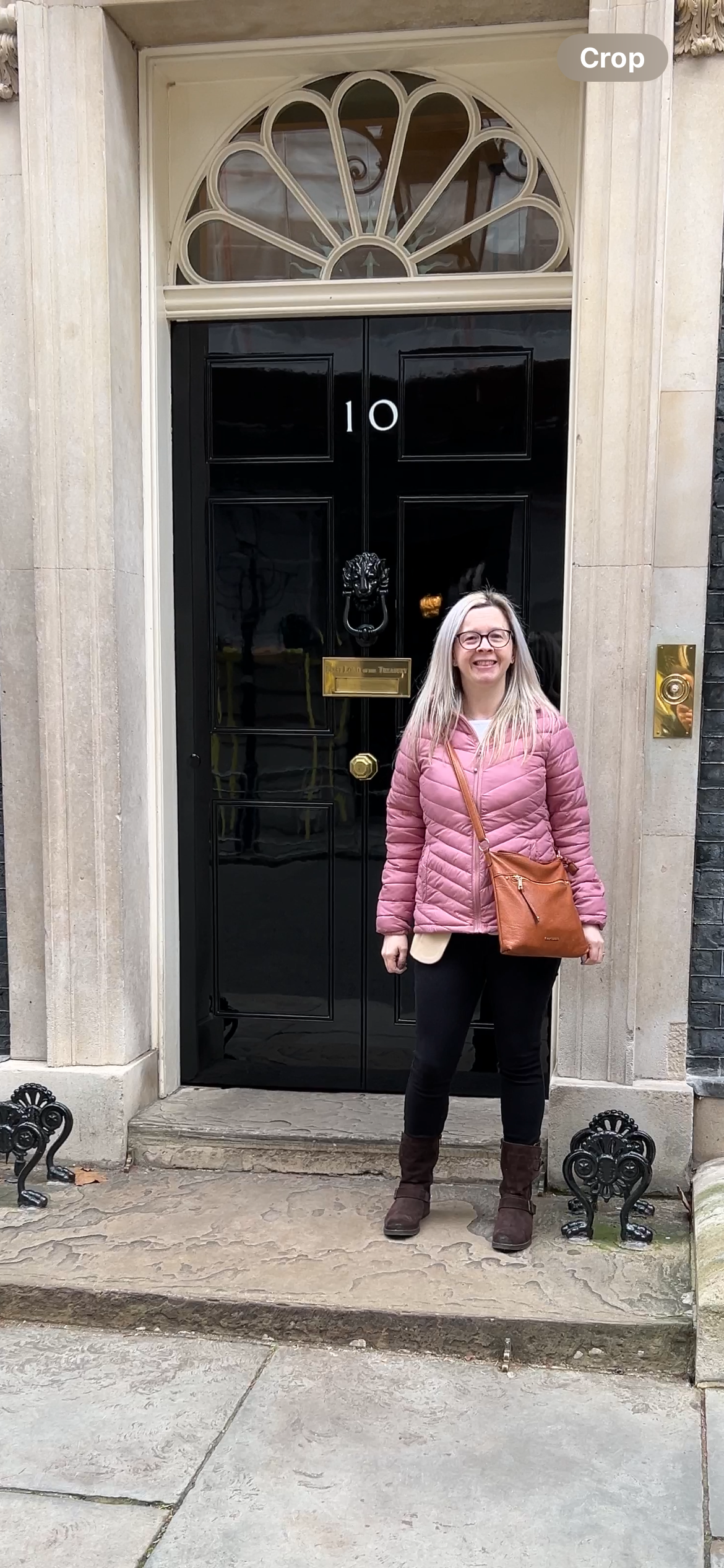 Hayley stood in front of 10 Downing Street, Hayley is wearing a pink coat, black trousers, brown jeans and a brown across body bag. Hayley is smiling at the camera.