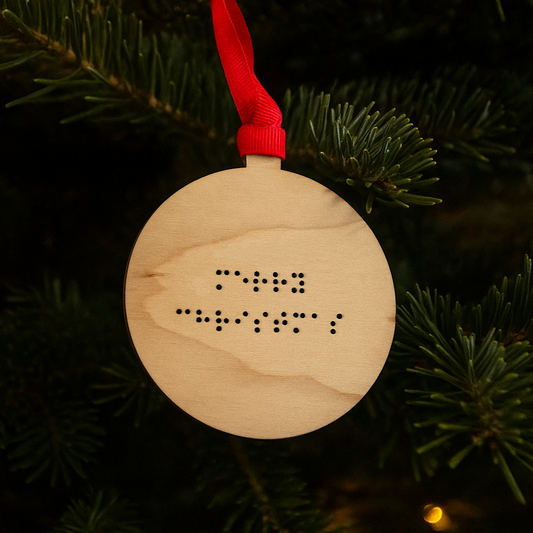 A round wooden Christmas tree decoration with Merry Christmas in lower case braille. The bauble has red ribbon and is hanging on a Christmas tree.