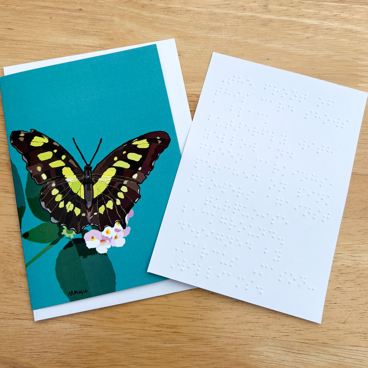 A greetings card showing a large black butterfly with yellow and green markings, perched on pink flowers against a turquoise background. There is a white braille note at the side.