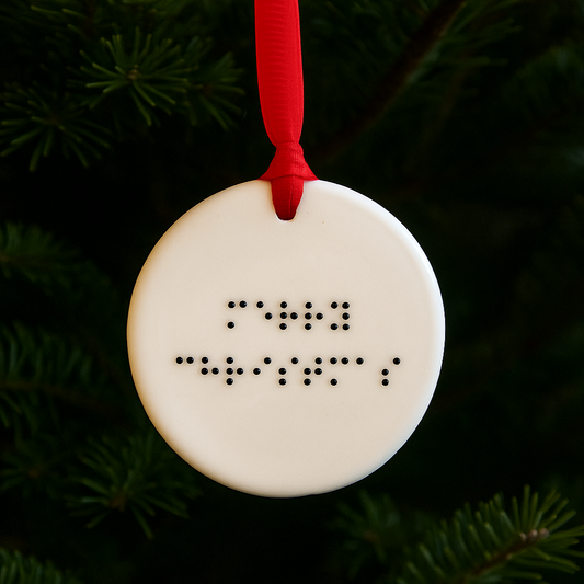 A circular ceramic Christmas tree decoration with merry christmas in grade one lower case braille, hanging on a tree with red ribbon.