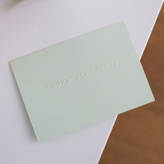 A pastel green card, lay flat on a table with congratulations written in lower case braille.