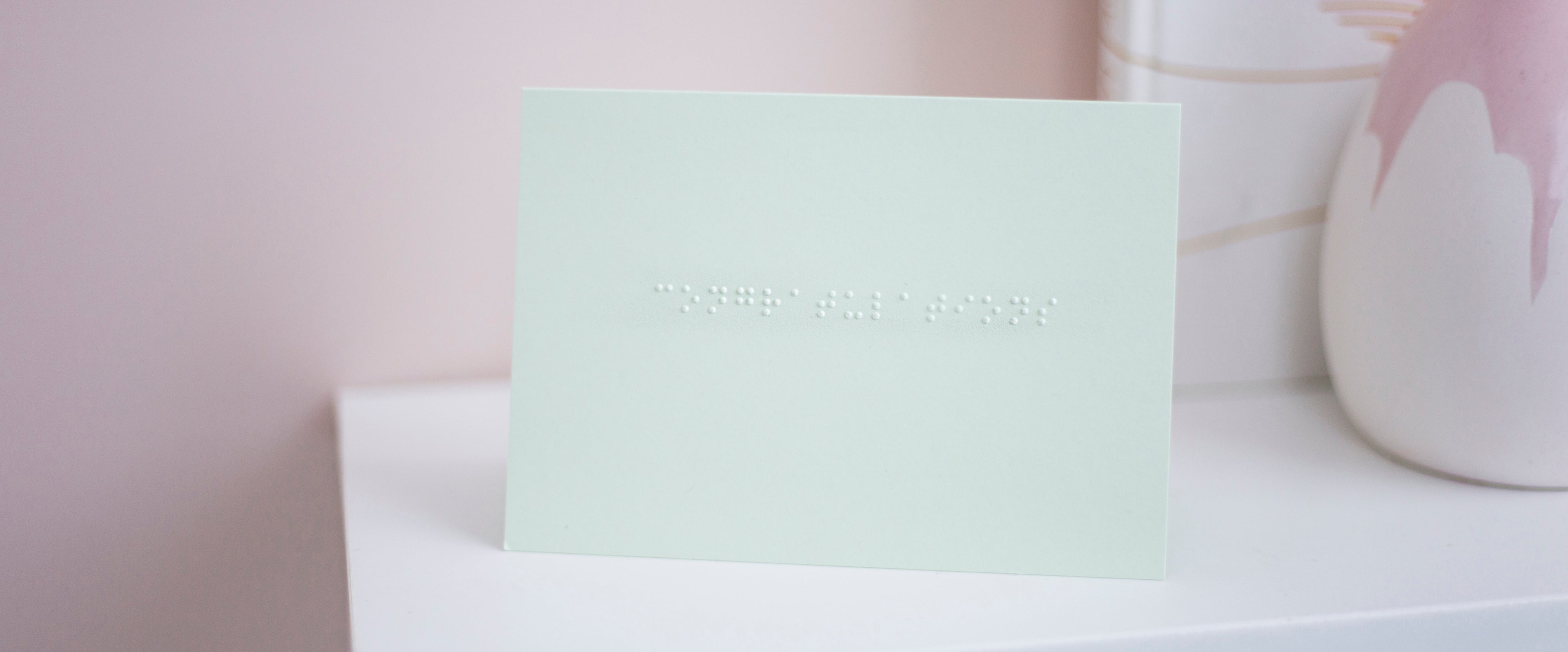 A pastel green card with congratulations written in lower case braille. The card is sat on a shelf with a vase just visible to the right. 