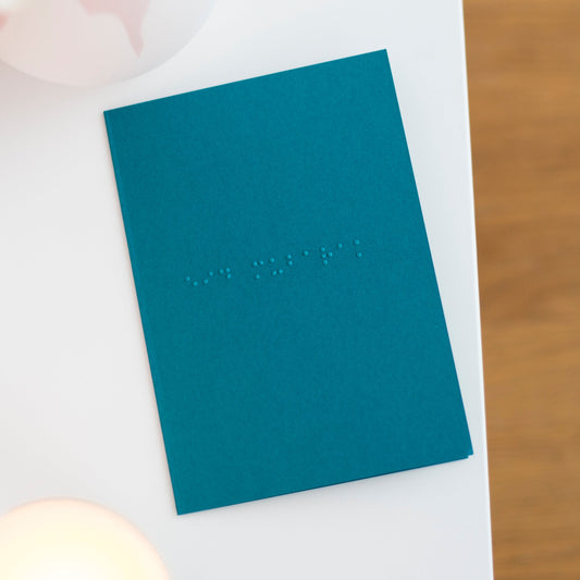 A teal coloured A6 greetings card with eid mubarak in lower case grade one braille.