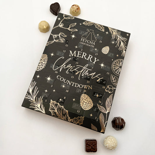 An advent calendar with a dark background and gold decorative elements. The text on the calendar reads "Fetcha Chocolates Merry Christmas Countdown." The calendar has 24 small, numbered doors and each door features braille label. The design includes festive illustrations such as pine cones, stars, holly leaves, and orange slices, all in a luxurious gold colour that contrasts against the black background. There are handcrafted chocolates dotted around.