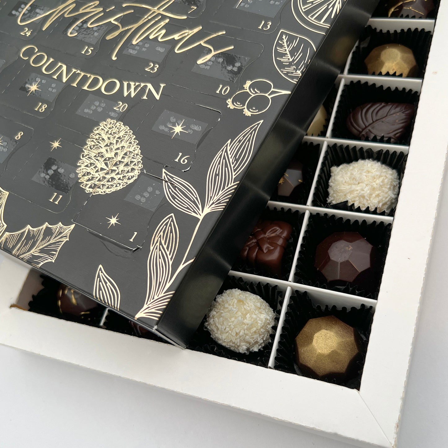A close up shot of the inside of the advent calendar, showing handcrafted luxury chocolates. The black advent calendar box is sat on top, at an angle.