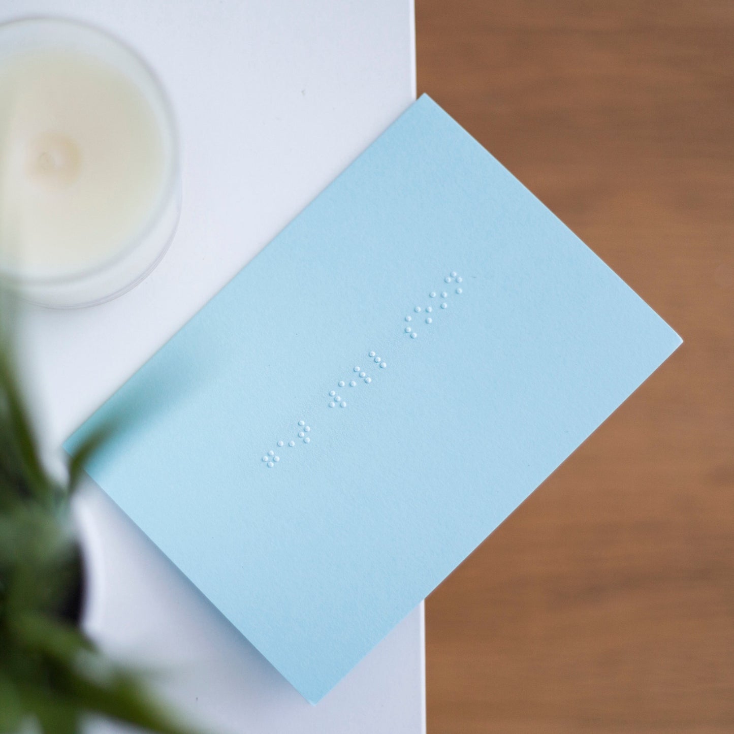 A pastel blue greetings card with get well soon written in lower case braille. There is a candle in the top left and a blurry plant in the bottom left.