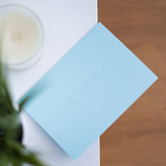 A pastel blue greetings card with get well soon written in lower case braille. There is a candle in the top left and a blurry plant in the bottom left.