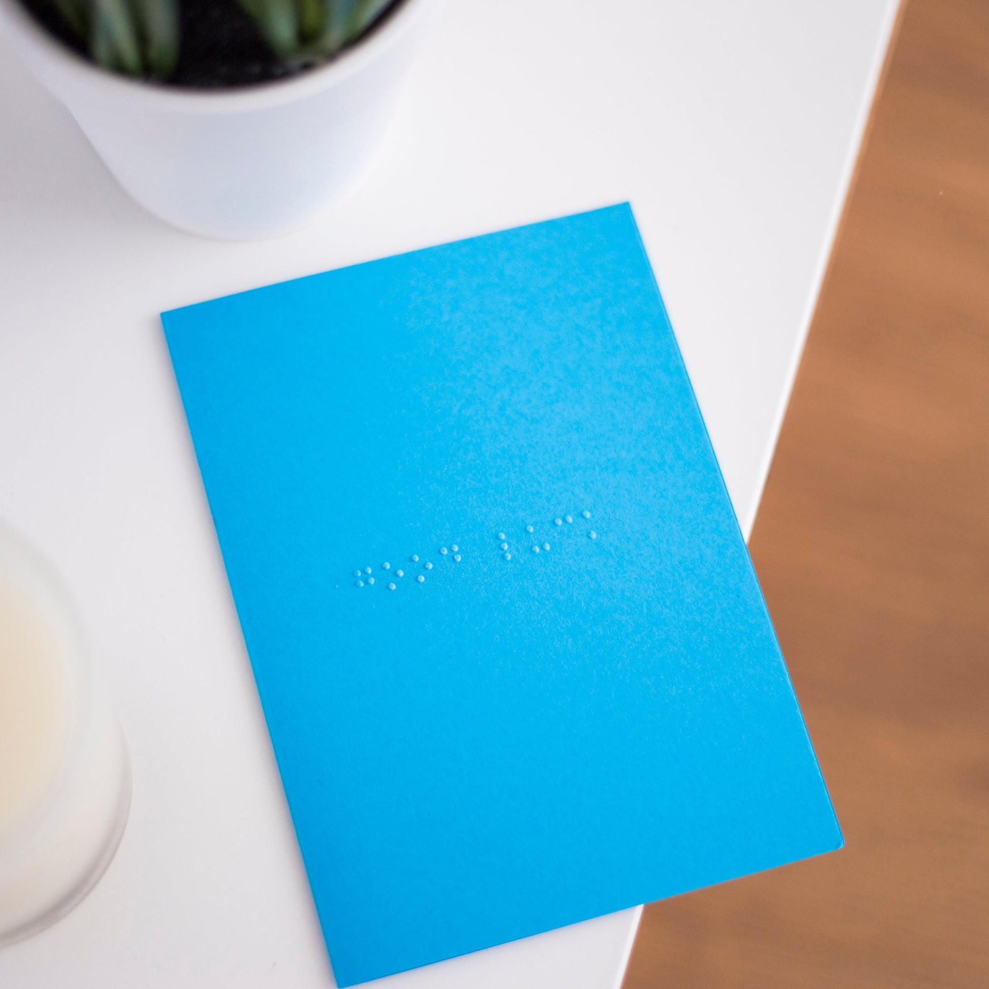 A vibrant blue card lay flat on a table with good luck written in lower case grade one braille. There is a candle in the bottom left and a blurry plant in the top left.