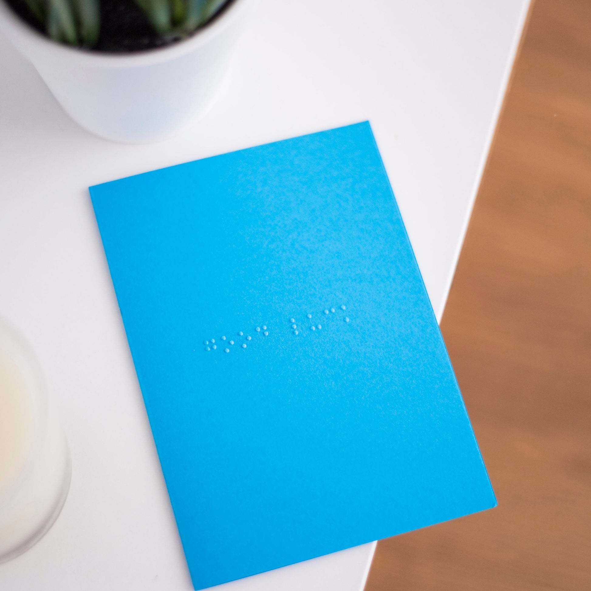 A vibrant blue card lay flat on a table with good luck written in lower case grade one braille. There is a candle in the bottom left and a blurry plant in the top left.