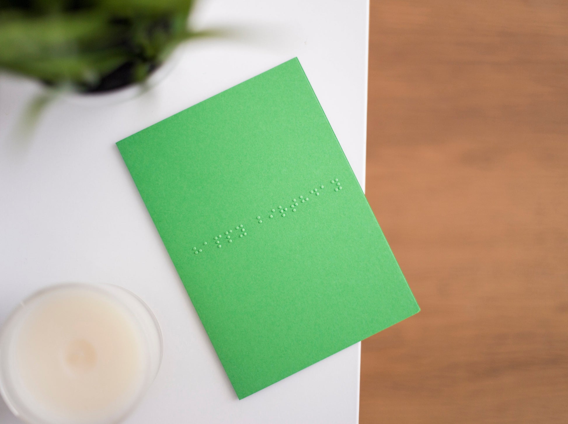 A vibrant green card lay flat on a table with happy birthday written in lower case grade one braille. There is a candle in the bottom left and a blurry plant in the top left.