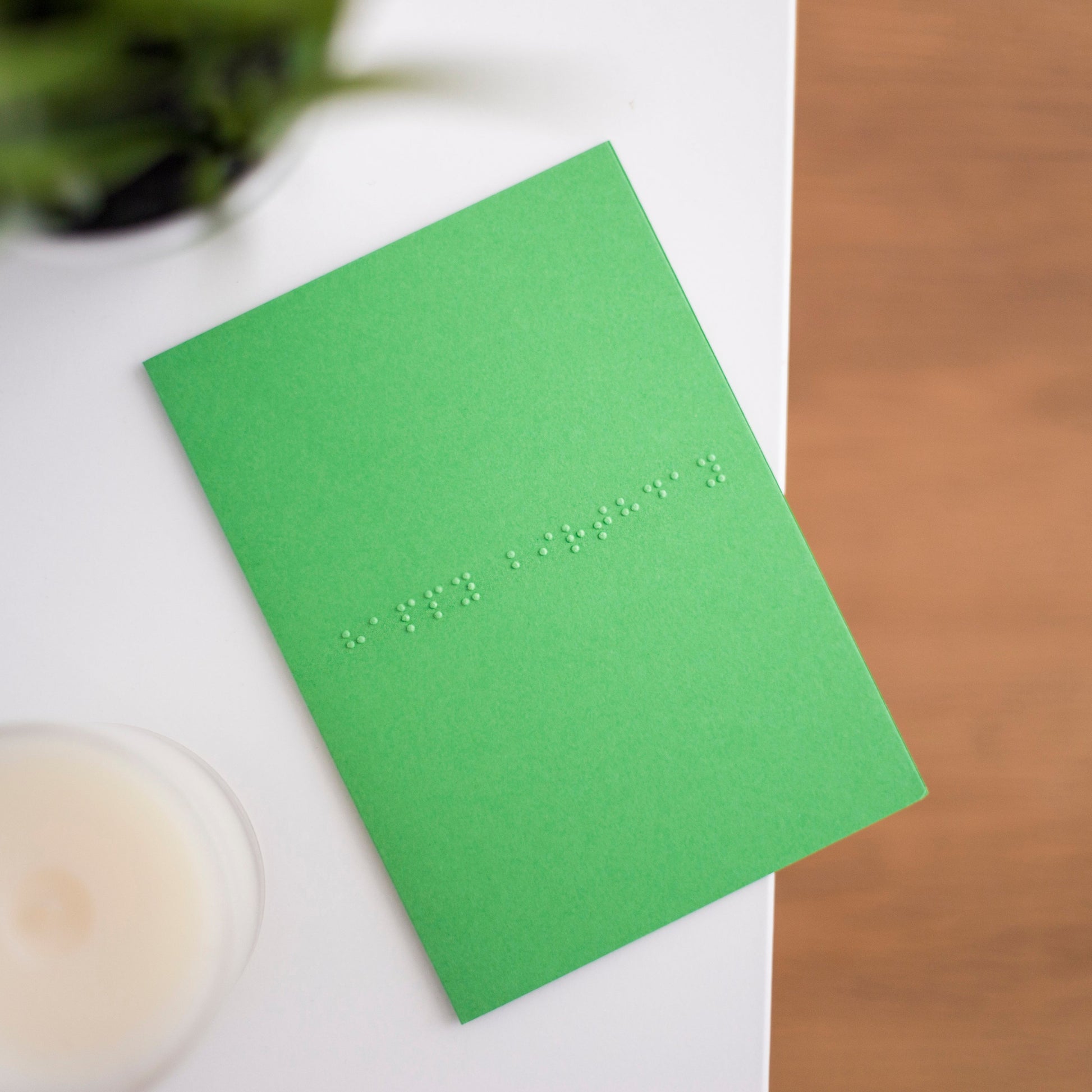 A vibrant green card lay flat on a table with happy birthday written in lower case grade one braille. There is a candle in the bottom left and a blurry plant in the top left.