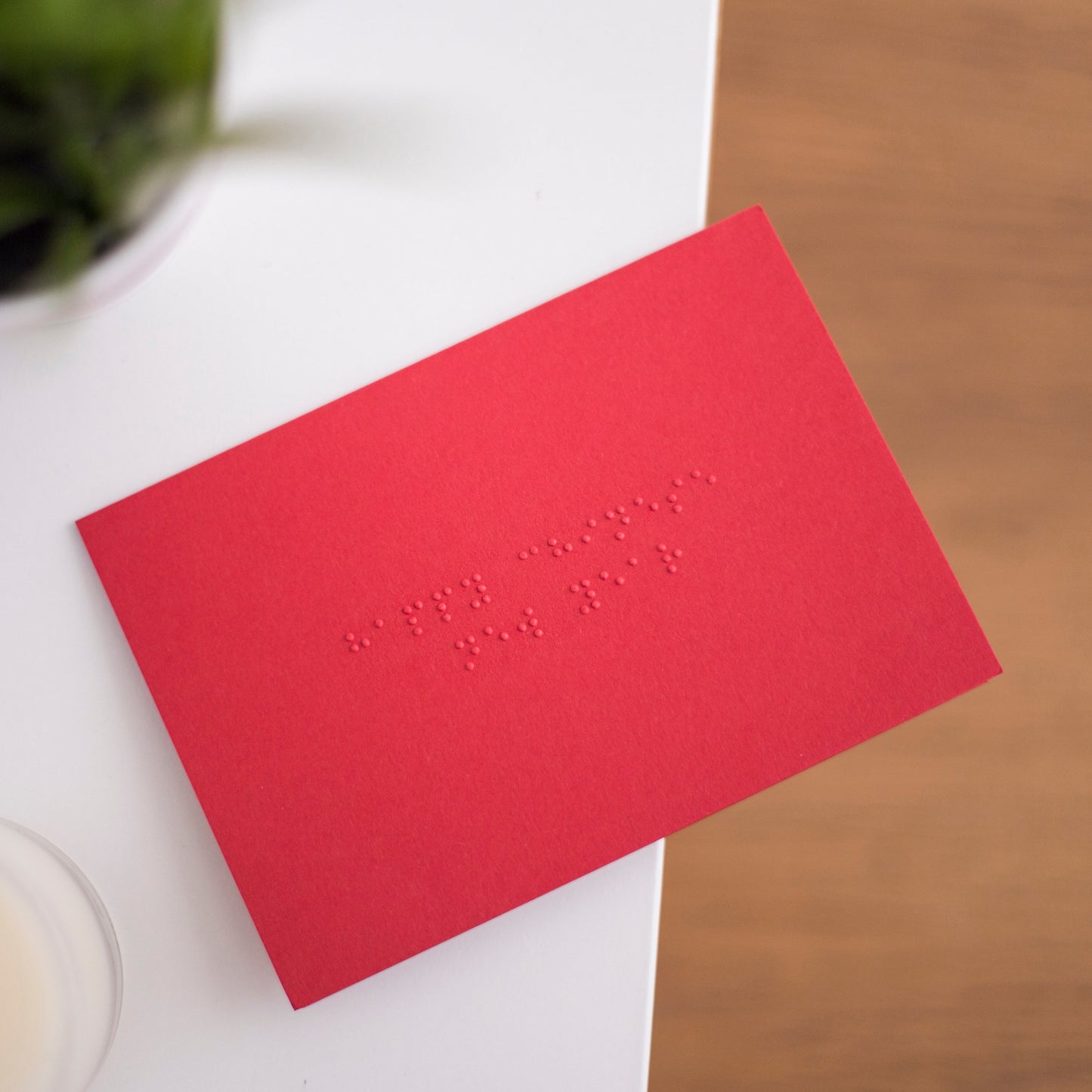 A vibrant red card lay flat on a table with happy Chinese new year written in lower case grade one braille. There is a candle in the bottom left and a blurry plant in the top left.