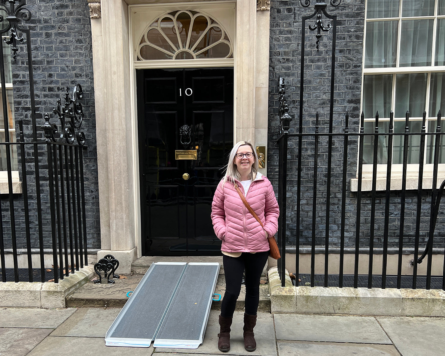 Hayley, a white blonde female, stood outside 10 Downing Street, there is a ramp leading up to the front door. Hayley is smiling at the camera.