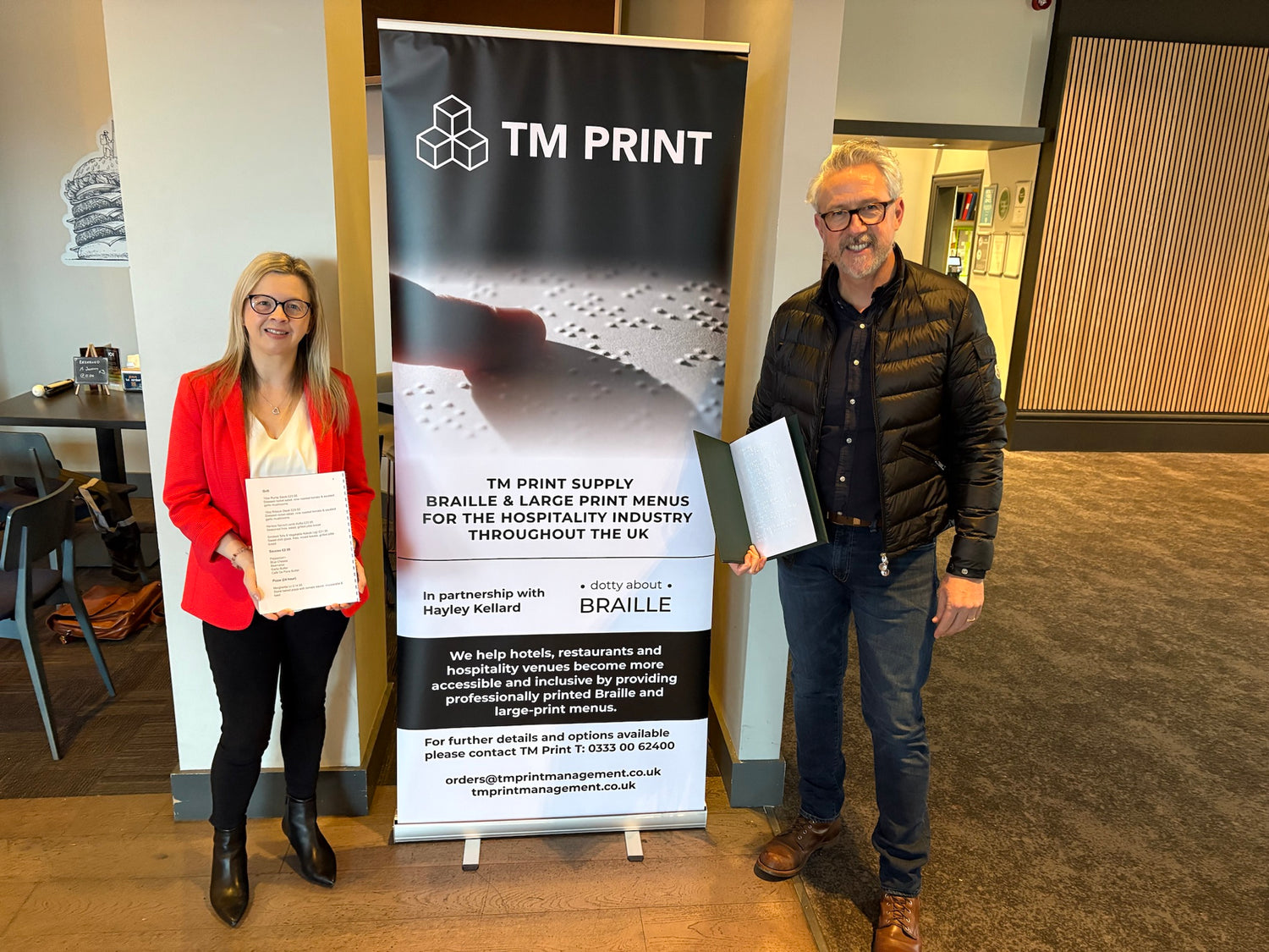 Hayley from Dotty About Braille stood with Glen from TM Print in front of a roller banner which advertises the braille and large print menus, they are each holding a menu.