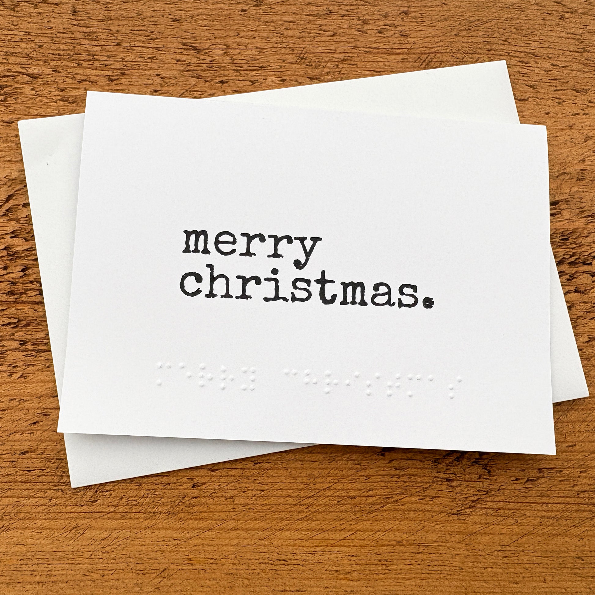 A white A6 card with Merry Christmas written in black typewriter style font with the same in braille beneath the printed text.
