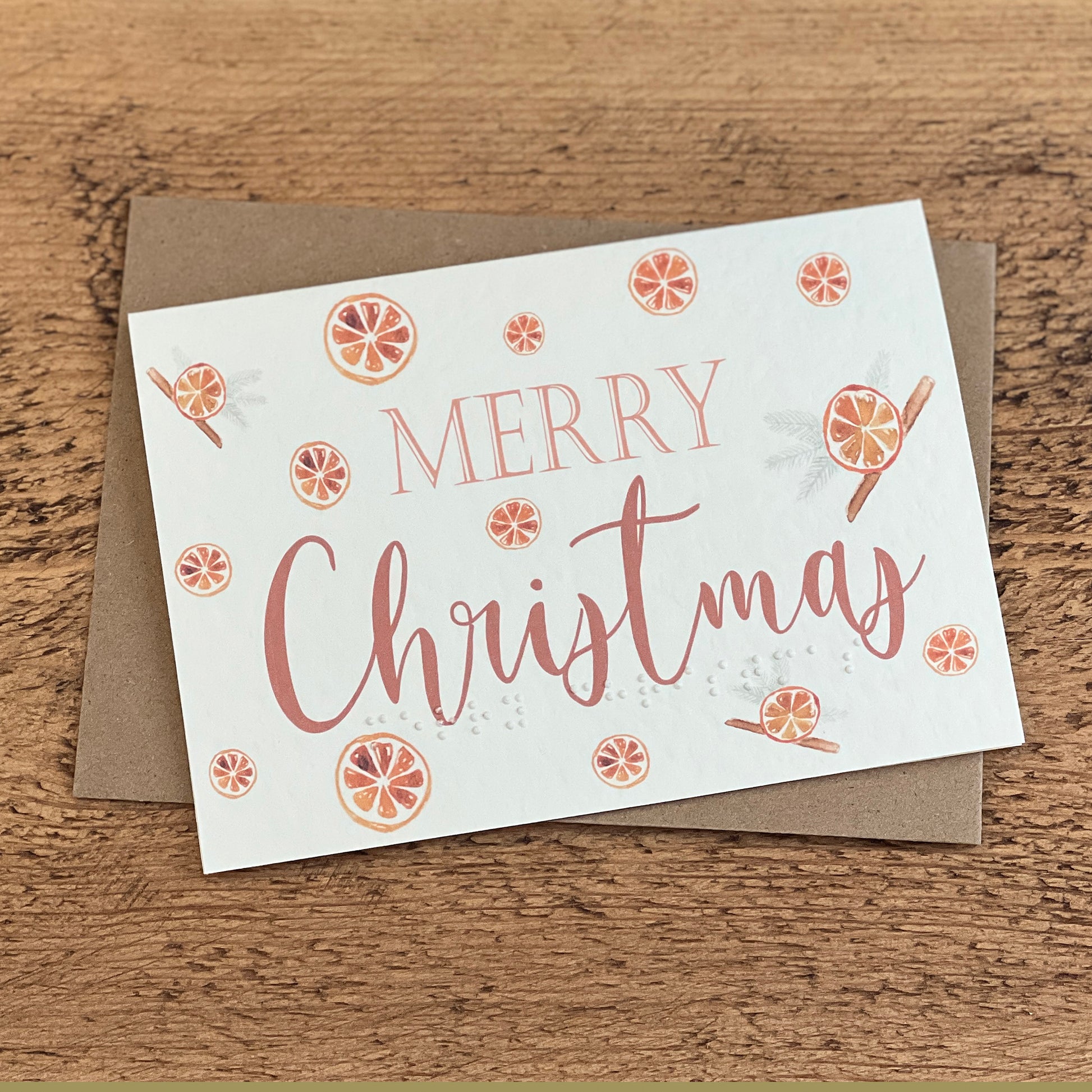 A textured white card with Merry Christmas written in a red font with citrus drawings around the card, the same text is written in braille along the bottom.