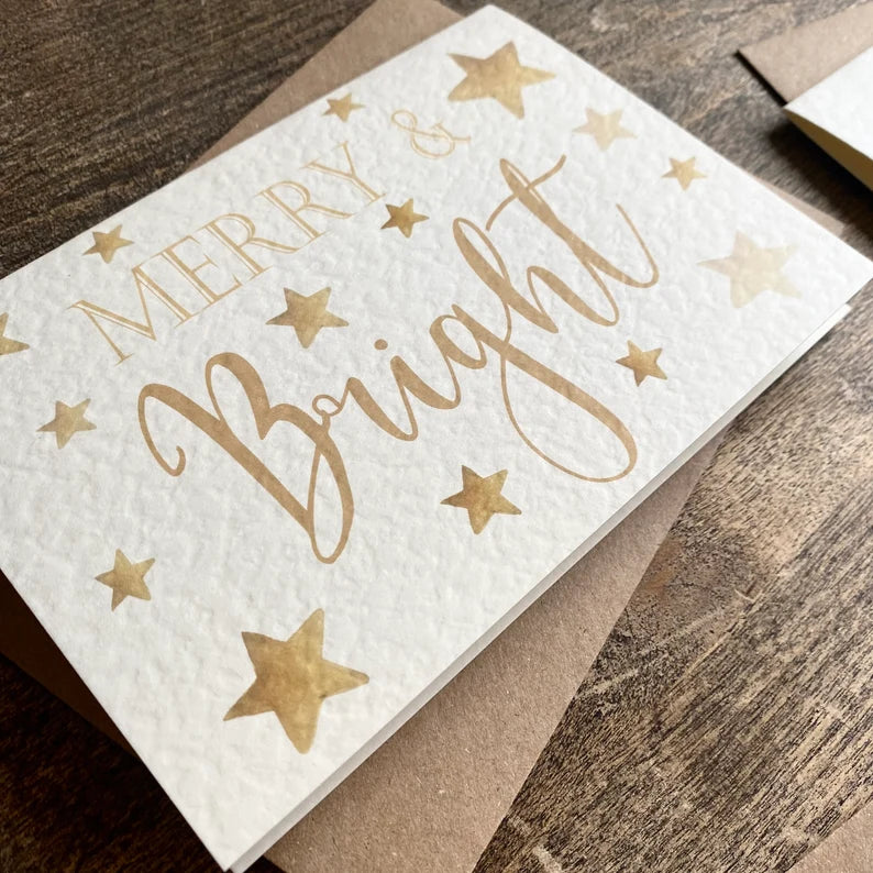 A textured white card with Merry & Bright written in a gold font with stars around the card.