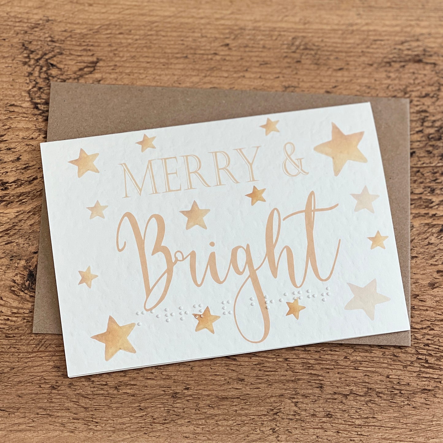 A textured white card with Merry & Bright written in a gold font with stars around the card, the same text is written in braille along the bottom.