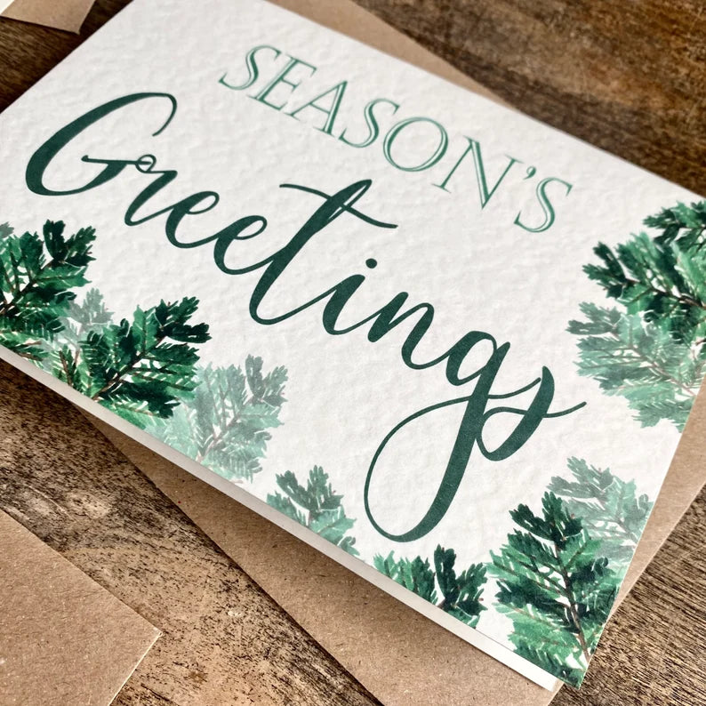 A textured white card with Season's Greetings written in a green font with foliage around the card.