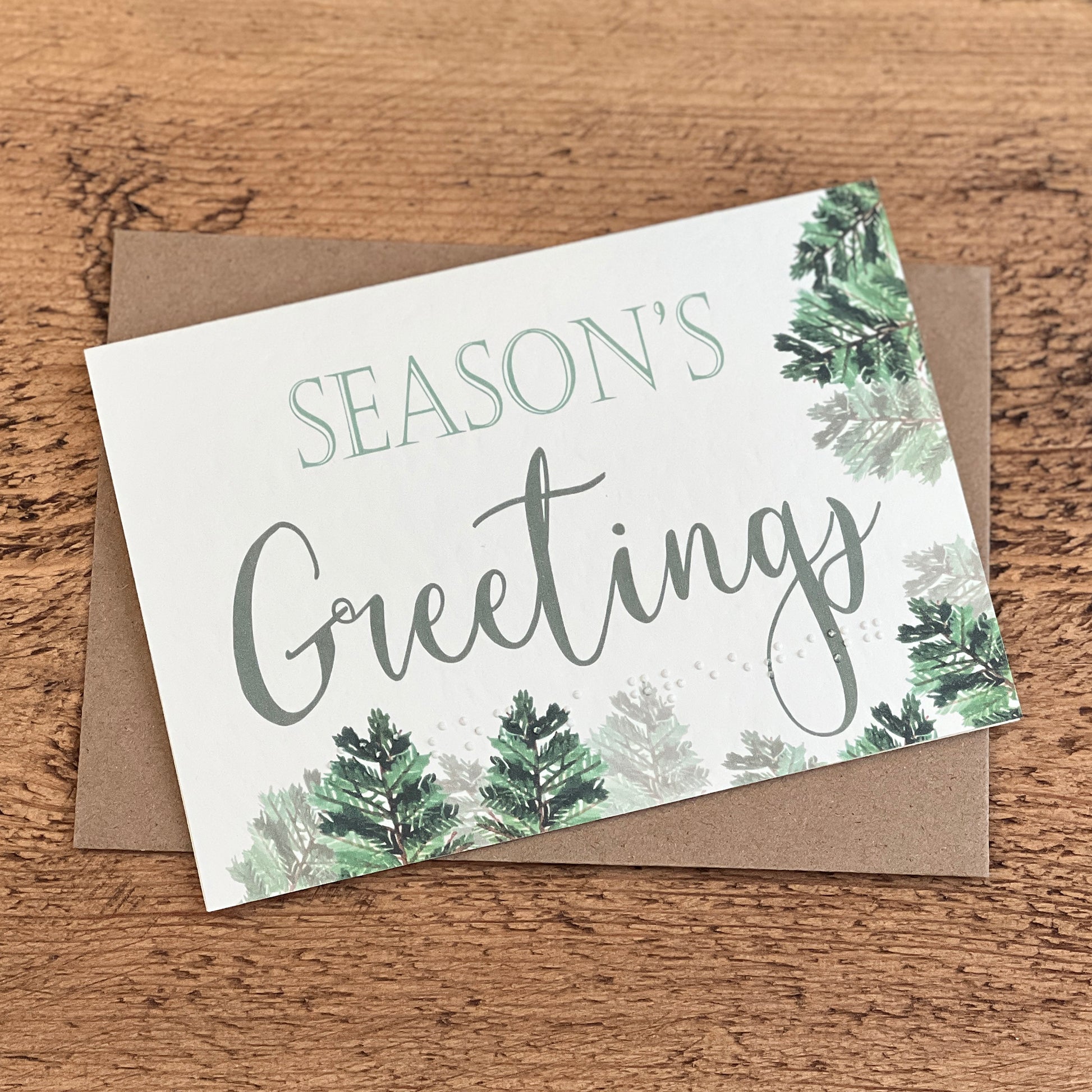 A textured white card with Season's Greetings written in a green font with foliage around the card, the same text is written in braille along the bottom.