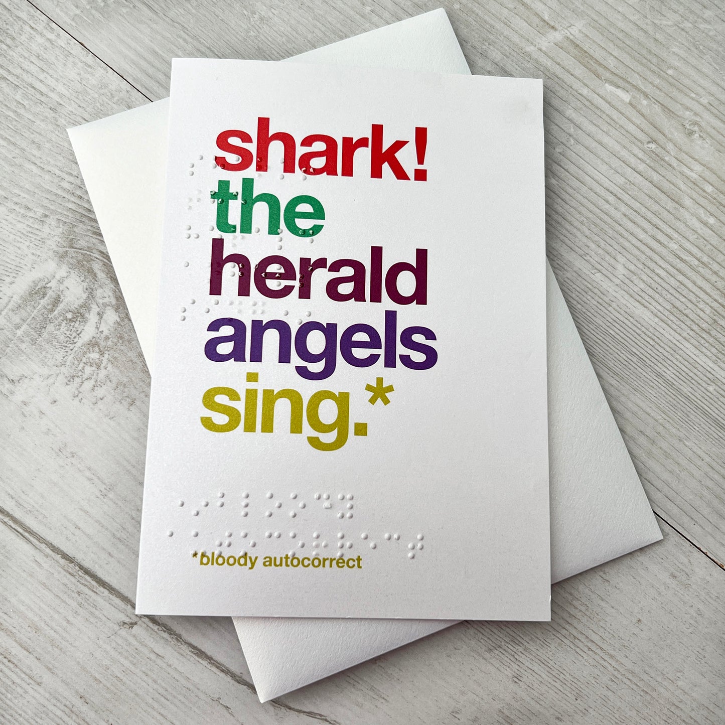 A white card with colourful text saying shark! the herald angels sing.* *bloody autocorrect with braille saying the same.