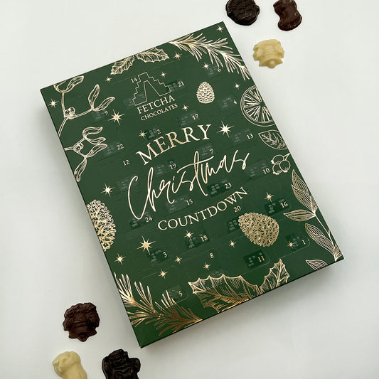 An advent calendar with a green background and gold decorative elements. The text on the calendar reads "Fetcha Chocolates Merry Christmas Countdown." The calendar has 24 small, numbered doors and each door features braille label. The design includes festive illustrations such as pine cones, stars, holly leaves, and orange slices, all in a luxurious gold colour that contrasts against the green background. There are milk, white and dark chocolates at the side.