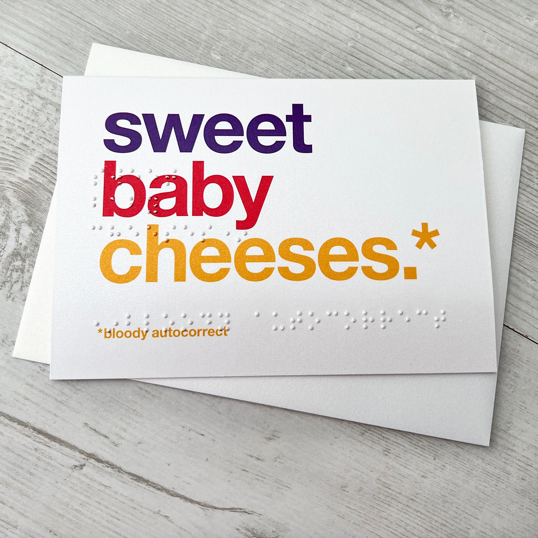 A white card with colourful text saying sweet baby cheeses.* *bloody autocorrect with braille saying the same.