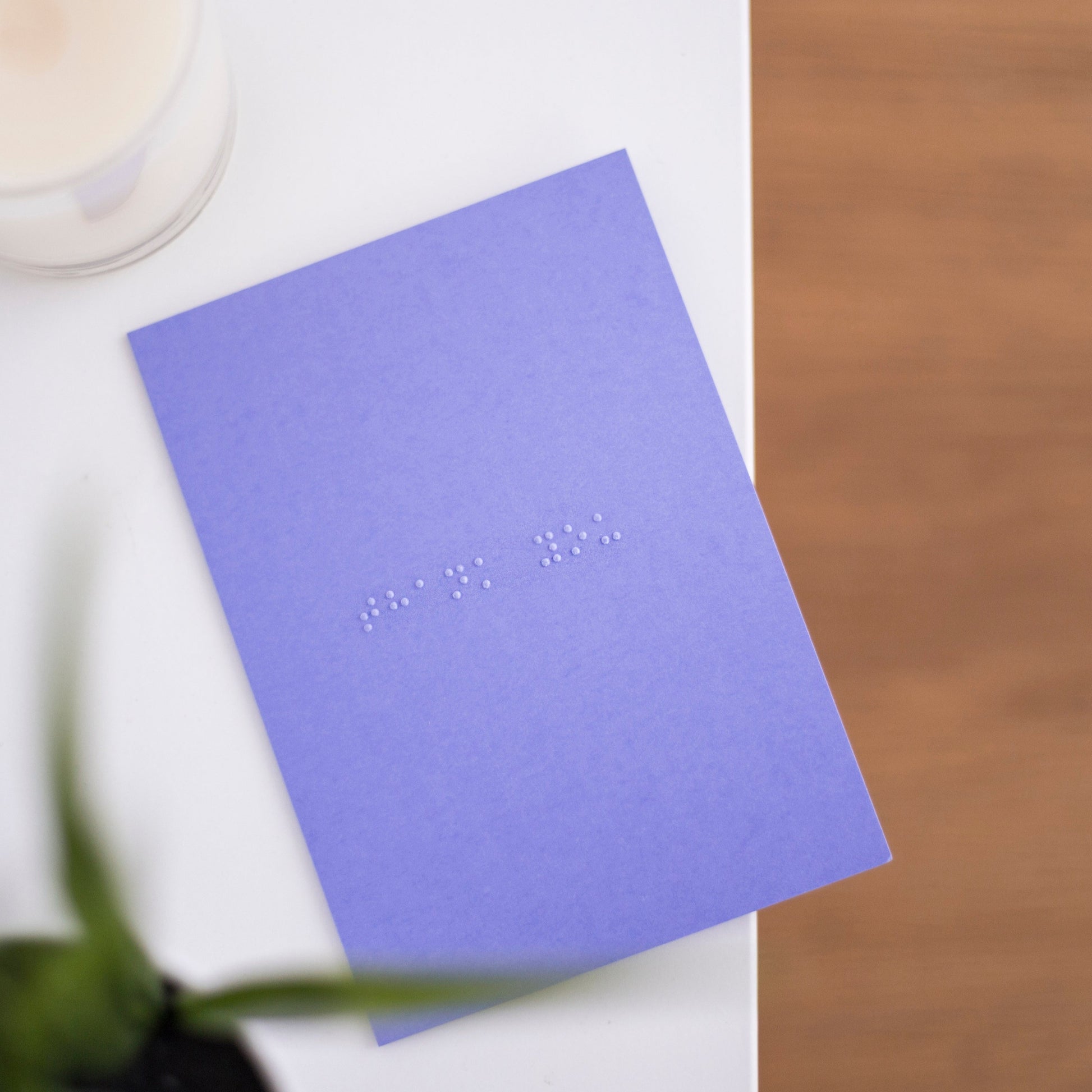 A vibrant purple card lay flat on a table with thank you written in lower case grade one braille. There is a candle in the top left and a blurry plant in the bottom left.