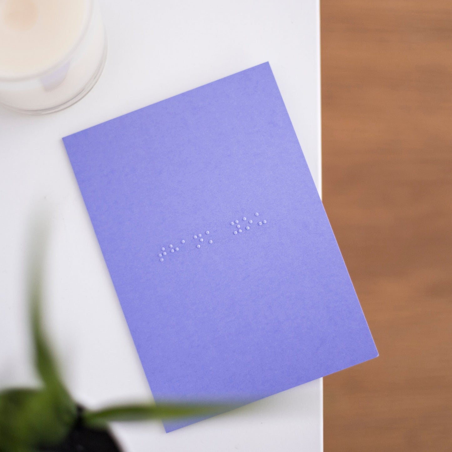 A vibrant purple card lay flat on a table with thank you written in lower case grade one braille. There is a candle in the top left and a blurry plant in the bottom left.