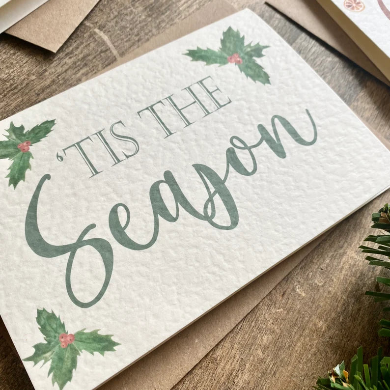 A textured white card with 'Tis The Season written in a green font with holly leaves around the card.