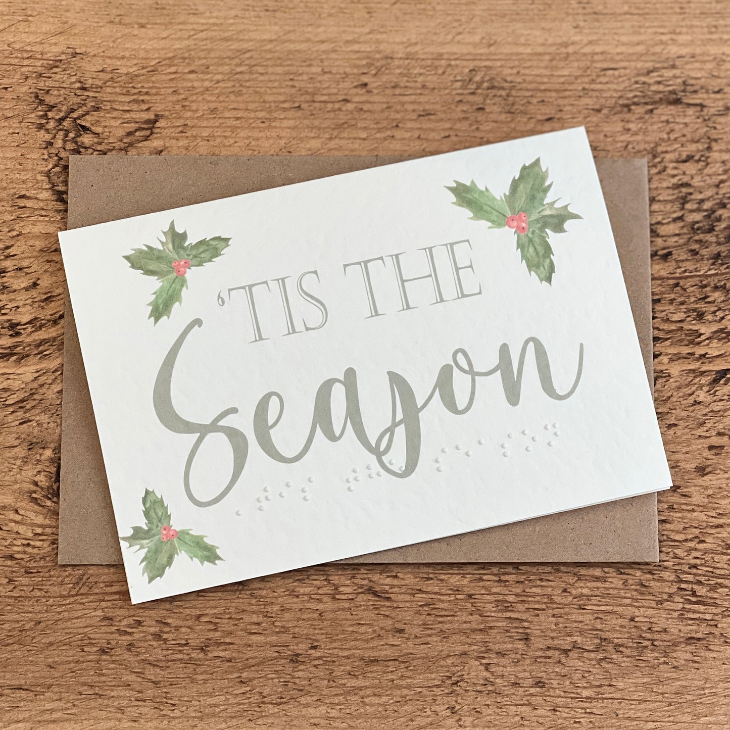 A textured white card with 'Tis The Season written in a green font with holly leaves around the card, the same text is written in braille along the bottom.