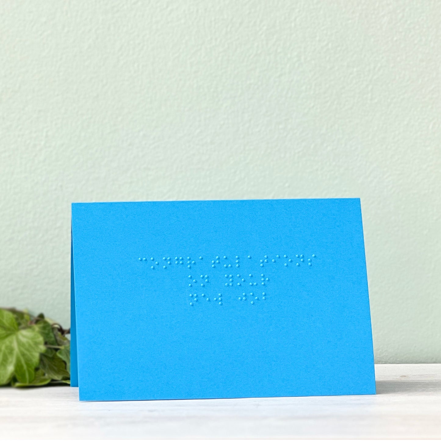 A vibrant blue card with "congratulations on your new job" written in lower case braille. There is foliage to the left of the card.