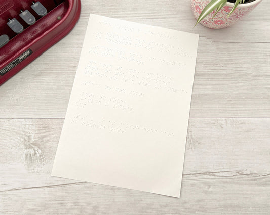 An A4 piece of white braille paper with a Perkins brailler to the left and a plant pot on the right.